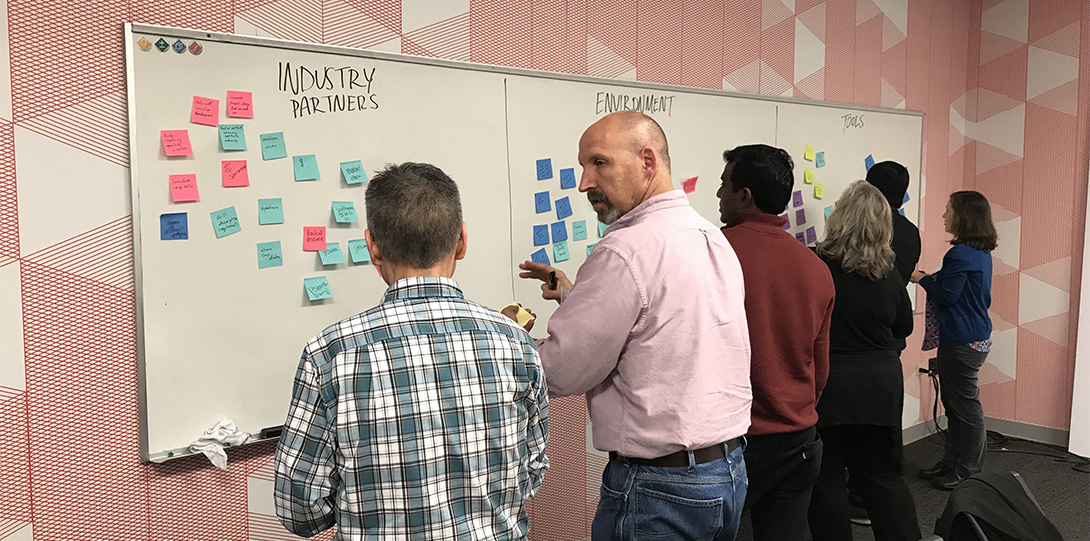 Six executive training participants working on a post it note clustering activity on a white board. Two participants are discussing what they are working on.