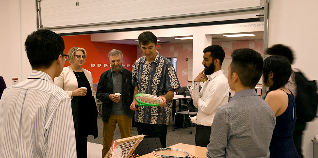 Students, faculty, and partners standing in a circle around a faculty member looking at a team's prototype of a tennis racket.