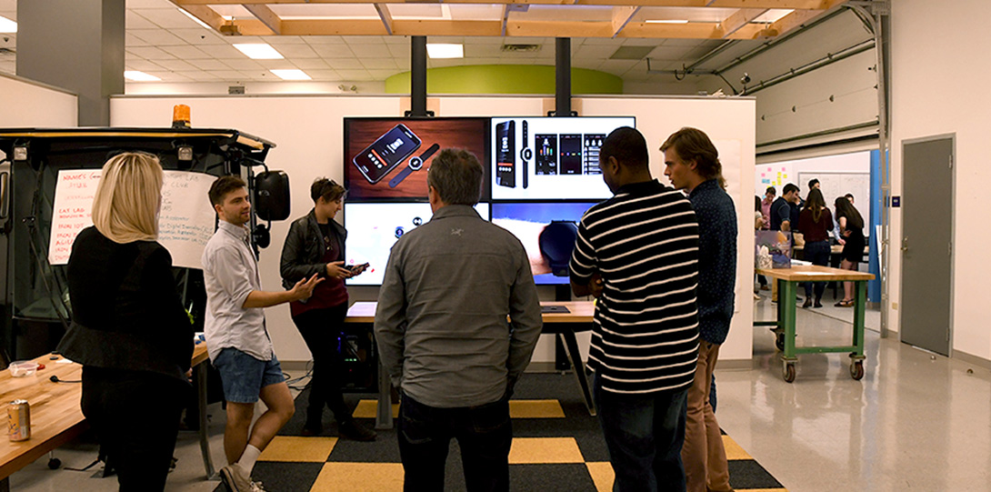 Students and partners standing around a television with four screen showing images of their prototype on a smartphone and smart watch.